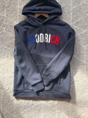 Mörkblå HOODRICH hoodie med brodyr - Snygg mörkblå hoodie från Hoodrich med broderad logga i blått, vitt och rött på bröstet. Klassisk huva med dragsko och stor magficka framtill. Perfekt för en chill och street-inspirerad stil.