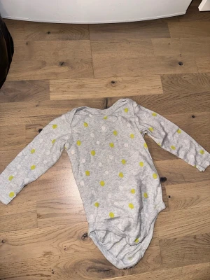 Grå långärmad body med prickar - Grå långärmad body från Lindex med vita och gula prickar. Mjuk och stretchig bomullskvalitet, perfekt för en bekväm vardagslook. Praktisk omlottöppning vid axlarna och tryckknappar i grenen för enkel på- och avklädning.