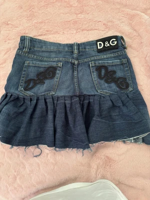 D&G blå volang jeanskjol - Cool blå jeanskjol från D&G med volang och råa kanter. Kjolen har klassiska fickor fram och bak, samt D&G-logga på bakfickorna och linningen. Perfekt för dig som gillar en edgy och trendig look.