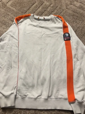 Vit sweatshirt med orange detalj PJS - Säljer en vit sweatshirt från Parajumpers med en bred orange rand längs axeln och ärmen samt ett PJS Anchorage-märke på bröstet. Tröjan har rund hals och ribbade muddar. Perfekt för dig som gillar streetwear och snygga detaljer.