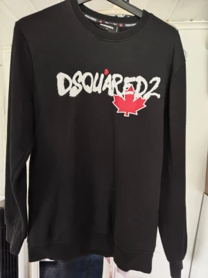 Svart sweatshirt från Dsquared2 - Cool svart sweatshirt från Dsquared2 med vit logga och röd lönnlövsgrafik på bröstet. Tröjan har rund halsringning och långa ärmar. Perfekt för dig som gillar streetwear och vill ha något trendigt i garderoben. Tyvärr finns de en liten fläck under armen baksida kolla kort Kan inte garantera äkthet 