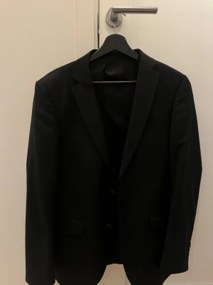 Svart klassisk kostym Jack & Jones  - Snygg svart kavaj med två knappar och klassisk krage. Fodrad insida med en diskret röd detalj längs sömmarna. Det som medkommer är svarta kostymbyxor och en väst också från Jack & Jones. Använd 2 gånger så i väldigt bra skick.
