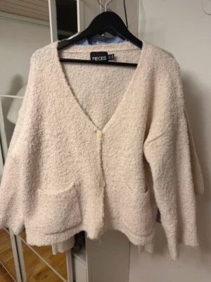 Ljusbeige kofta från Pieces - Mysig ljusbeige kofta från Pieces med två framfickor och v-ringning. Koftan har en fluffig, bouclé-liknande textur och stängs med två knappar framtill. Perfekt för lager-på-lager och chill dagar.