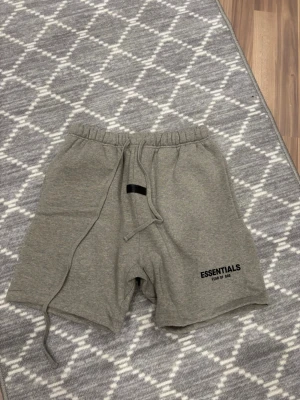 Grå Essentials shorts  - Säljer ett par grå shorts från Fear of God Essentials med elastisk midja och snörning. Shortsen har en avslappnad passform och logga tryckt på vänster ben. Perfekta för chill dagar och streetwear-stil.