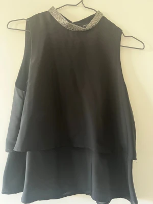 Svart ärmlös topp med glittrig krage - Snygg svart ärmlös topp från Zara med lager-på-lager-effekt och glittrig silverkrage. Perfekt för dig som vill sticka ut lite extra. Toppen har en lös passform och är tillverkad i ett mjukt, lätt material.