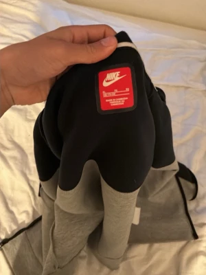 Nike tvåfärgad hoodie svart/grå - Snygg hoodie från Nike i svart och grått med dragkedja och vit logga på bröstet. Hoodien har huva och är tillverkad i mjukt bomullsmaterial. Perfekt för en avslappnad och sportig stil.