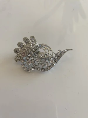 Silverfärgad broschyr med strass - En snygg  broschyr i silverfärg med glittrande strasstenar och ett dekorativt, blommigt mönster. . Materialet är metall och dekoren består av klara stenar.