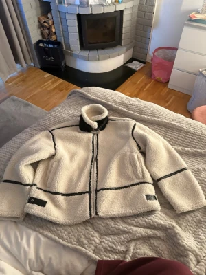 Vit teddyjacka med svarta detaljer - Säljer en fluffig vit teddyjacka med svarta detaljer och dragkedja framtill. Jackan har hög krage, svarta kantband och två fickor på sidorna. Perfekt för kalla dagar när du vill vara både varm och snygg.