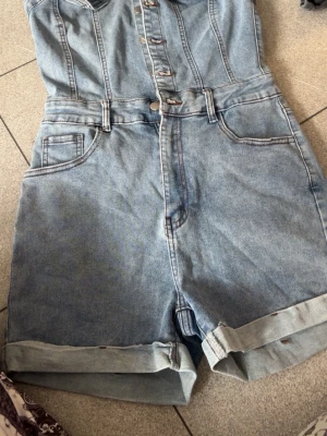 Ljusblå jeansbyxdress med shorts - Snygg ljusblå jeansbyxdress med knappar framtill och uppvikta benslut. Modellen har klassiska fickor fram och bak samt bälteshällor i midjan. Perfekt för en avslappnad och trendig look. Materialet är denim i bomull.