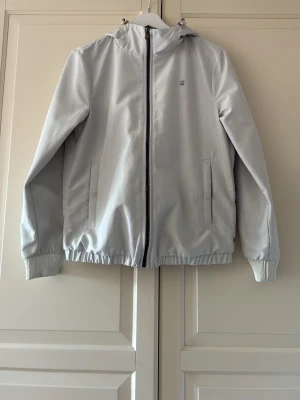 Ljusgrå vindjacka från G-Star RAW - Snygg ljusgrå vindjacka från G-Star RAW med dragkedja framtill, två fickor och ribbade muddar vid ärmslut och midja. Jackan har en minimalistisk design med liten logotyp på bröstet och huva. Storlek M, men är liten i storlek, skulle säga att den är mer som S. Finns en liten fläck på jackan och jag har behandlat den, lyckades inte få bort resten (se bild). Annars är jackan i fint skick. 
