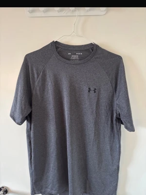 Grå tech t-shirt från Under Armour - Snygg grå tech t-shirt från Under Armour med klassisk rund hals och diskret logga på bröstet. Modellen är loose fit och tillverkad i ett mjukt syntetmaterial som andas, perfekt för träning eller chill dagar.