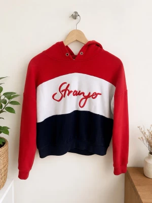 Röd, vit och marinblå hoodie Stranger - Snygg hoodie i rött, vitt och marinblått med texten 'Stranger' broderad på bröstet. Hoodien har huva med dragsko och är tillverkad i mjukt sweatshirtmaterial. Perfekt för en avslappnad och trendig look.