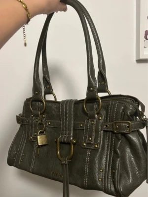 Vintage looking handväska från Bdg - Snygg mörkgrön handväska från Bdg med dubbla handtag, dekorativa nitar och mässingsfärgade metalldetaljer. Väskan har ett hänglås med nyckel, vita sömmar och ett spänne framtill. Måtten: 36 bredd och 21 höjd, rymmer mycket. Använd flera gånger men i bra skick 