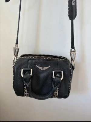 Svart axelväska från Zadig & Voltaire - Cool svart axelväska i läder från Zadig & Voltaire med silverfärgade detaljer och nitar längs kanterna. Väskan har justerbar axelrem med logga, dubbla handtag och en snygg vinge i metall framtill. Den är i super bra skick endast lite rost på metallen annars super fin! Kvitto finns! Kom med prisförslag 