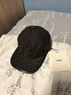 Svart Fendi keps med FF-mönster - Snygg svart keps från Fendi med det klassiska FF-logomönstret över hela kepsen. Kepsen har böjd skärm och justerbart band bak för perfekt passform. Tillverkad i ett slitstarkt tyg som känns lyxigt och trendigt. Perfekt accessoar för dig som gillar designerplagg.