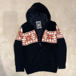 Fodrad Cardigan - Säljer en svart fodrad cardigan med ett snyggt rött och vitt mönster över bröstet och ryggen. Tröjan har dragkedja och ribbstickade muddar, perfekt för kyliga dagar. Den har en quiltad insida för extra värme. Vid minsta fråga eller fundering hör av er🤝