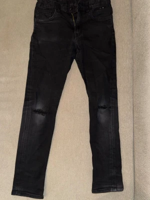 Svarta skinny jeans med slitningar - Svarta skinny jeans med slitna detaljer vid knäna och klassisk femficksdesign. Jeansen har en något tvättad look och smal passform som sitter tajt längs benen. Perfekta för en trendig och avslappnad stil.