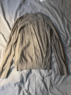 Beige tunn stickad tröja - En stilren beige stickad tröja med rund halsringning och långa ärmar. Tröjan är tunn och har ribbade muddar vid ärmslut och nederkant. Perfekt för lager-på-lager och enkel att matcha med olika outfits.