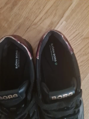 Svarta Björn Borg sneakers med metallicdetalj - Snygga svarta sneakers från Björn Borg med metallicdetalj i vinrött på hälen och guldfärgad logga på sidan. Skorna har klassisk låg profil, svarta snören och ovandel i läderimitation. Perfekta för en stilren och modern look.