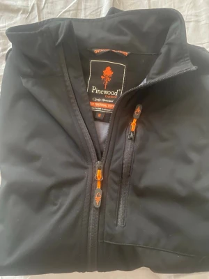 Svart vindjacka från Pinewood - Svart vindjacka från Pinewood med orange detaljer på dragkedjorna. Jackan har hög krage, bröstficka med dragkedja och är tillverkad i ett slitstarkt material som passar perfekt för utomhusaktiviteter. Snygg och funktionell design.