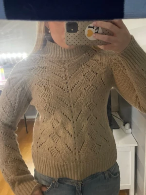 Beige stickad tröja med mönster - Säljer en beige stickad tröja med snyggt hålmönster och hög krage. Tröjan har långa ärmar och ribbade muddar vid ärmslut och nederkant. Perfekt för att hålla sig varm och stilren under kalla dagar.