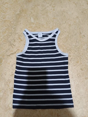 Randig linnetopp från Lindex - Snygg randig linnetopp från Lindex i marinblått och vitt. Toppen är ärmlös med ribbade kanter och har en klassisk, enkel design som passar perfekt till sommaren. Tillverkad i mjuk bomull för skön känsla hela dagen.