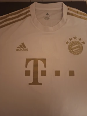 Vit Bayern München tröja Adidas - Snygg vit matchtröja från Bayern München med gulddetaljer och Adidas-logga. Tröjan har klubbmärke, sponsortryck och tre guldiga ränder på axlarna. På ärmen finns Bundesliga-märke för säsongen 2021/2022. Perfekt för fotbollsfans!