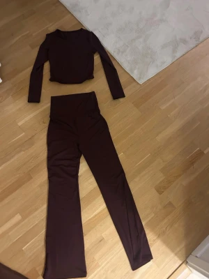 Vinrött set med långärmad topp - Snyggt tvådelat set i mörkbrun färg med långärmad, figurnära topp och matchande yoga pants med utsvängda ben. Setet är gjort i ett stretchigt och mjukt material som sitter bekvämt på kroppen. Perfekt för en trendig och avslappnad look.