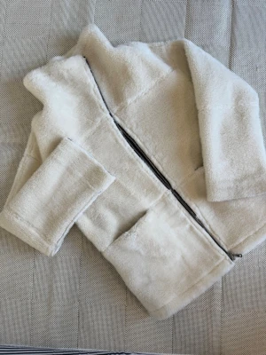 Krämvit teddyjacka med dragkedja - Supermjuk teddyjacka i krämvit färg med hel dragkedja framtill och två stora fickor. Jackan har en boxig passform och är tillverkad i fluffigt teddy-material som ger en mysig känsla. Perfekt för kalla dagar när du vill vara både varm och stylish.