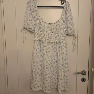 ny! Vit blommig klänning med puffärm - Söt vit klänning med ljusblått blommönster och puffärmar. Klänningen har knytband vid ärmarna och en smickrande v-ringning. Perfekt för varma dagar och har en romantisk vibe.prislapp kvar