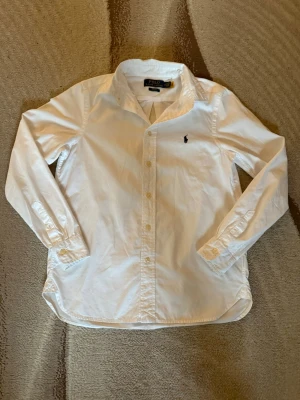Vit skjorta från Polo Ralph Lauren - Klassisk vit skjorta från Polo Ralph Lauren med broderad logga på bröstet. Skjortan har lång ärm, knappar framtill och är tillverkad i bomull. Perfekt för en clean och stilren look. Storlek är year 16 men passar som en XS