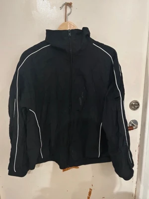 Svart vindjacka med vita detaljer - Svart vindjacka med dragkedja framtill och vita ränder längs ärmar och sidor. Jackan har hög krage och en sportig look, perfekt för blåsiga dagar. Materialet är tunt och lätt, vilket gör den smidig att bära. Passar dig som gillar enkel och stilren design. Har en liten fläck framtill som är synlig på bilden.