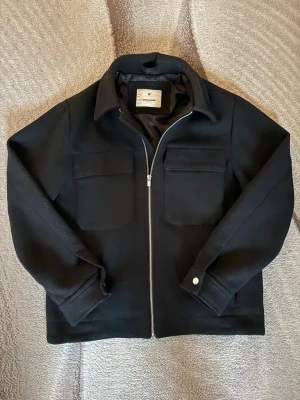 Svart overshirt från Jack & Jones - Stilren svart overshirt från Jack & Jones med två stora bröstfickor, dragkedja framtill och klassisk krage. Overshirten är i ett kraftigare tyg som ger en snygg och clean look. Perfekt att slänga över en hoodie eller t-shirt.