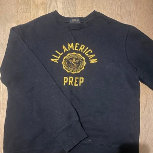 Mörkblå sweatshirt Polo Ralph Lauren - Mörkblå sweatshirt från Polo Ralph Lauren med gul text och emblem på bröstet där det står 'All American Prep'. Klassisk rund halsringning och ribbade muddar. Perfekt för en avslappnad och sportig stil. Funkar för barn i 9-11 års åldern