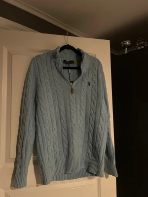 Ljusblå kabelstickad tröja Polo Ralph Lauren - Ljusblå kabelstickad tröja från Polo Ralph Lauren med halv dragkedja och ribbade muddar. Tröjan har klassisk broderad logga på bröstet och hög krage. Perfekt för lager-på-lager och en chill stil.