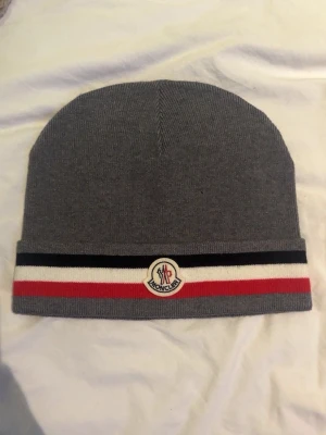 Grå Moncler Mössa  - Snygg grå mössa från Moncler med klassisk logga framtill. Mössan har ribbstickad struktur och en randig kant i mörkblått, vitt och rött. Perfekt för att hålla värmen och ge en stilren look. Materialet är mjukt och stretchigt, vilket gör den bekväm att bära. Den är använd typ 2 gånger och därmed i nyskick.