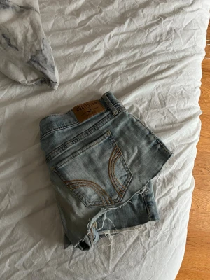 Ljusblå jeansshorts från Hollister - Snygga ljusblå jeansshorts från Hollister med råa kanter och klassiska fem fickor. Shortsen har låg midja och slitna detaljer för en avslappnad look. Perfekta för varma dagar och enkel att matcha med allt.