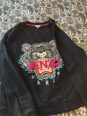 Svart Kenzo sweatshirt med tiger - Svart sweatshirt från Kenzo med ikonisk broderad tiger och färgglad logga på bröstet. Rund halsringning och ribbade muddar. Stora broderade detaljer i rosa, turkos, beige och vitt. Perfekt statement-plagg för dig som gillar streetwear.