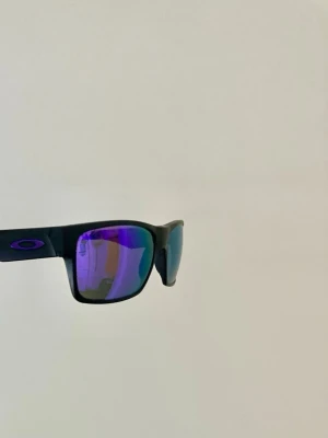 Oakley Twoface solglasögon  - Snygga svarta solglasögon från Oakley med rektangulära bågar och spegelglas i lila och blå toner. Klassisk Oakley-logga på sidan. 