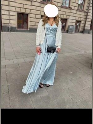 Ljusblå långklänning i satin Ivory Field  - Elegant ljusblå långklänning i glansig satin med omlottliv, v-ringning och öppen rygg med snörning. Klänningen har tunna axelband och ett svepande fall som ger en lyxig känsla. Perfekt för speciella tillfällen.