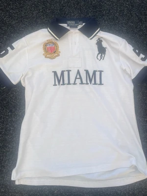 Polo Ralph Lauren skjorta MIAMI - Vit pikéskjorta från Polo Ralph Lauren med mörkblå detaljer, stor logga på bröstet, broderad patch och texten MIAMI framtill. Nummer 5 på ryggen och ärmarna. Klassisk krage och knappar framtill.