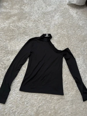 Svart oneshoulder långärmad topp - Trendig svart långärmad topp med asymmetrisk design och oneshoulder-skärning. Hög krage på ena sidan ger en cool och modern look. Perfekt att styla med jeans eller kjol för en edgy vibe.