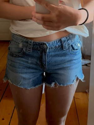 Blå jeansshorts med fransig kant - Snygga blå jeansshorts med fransig, rå kant och slitna detaljer. Klassisk femficksmodell med knapp och dragkedja framtill samt bälteshällor. Bakfickor med lock och knapp. Perfekta för sommaren och ger en avslappnad look. Storlek barn 170 men passar perfekt på mig som är en S!