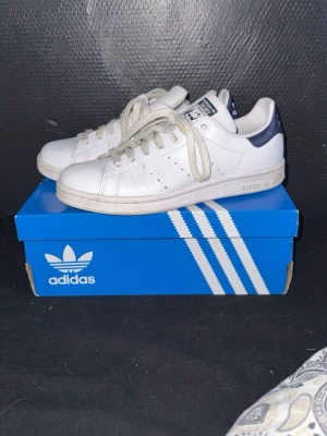Adidas Stan Smith vita sneakers - Klassiska vita Adidas Stan Smith sneakers med mörkblå detalj på hälen . Skorna har lågt skaft, perforerade ränder på sidorna och är tillverkade i läder med gummisula. Perfekt för en clean och tidlös stil. Andvända 1-2 ggr nypris ca 1400kr