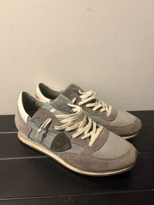 Grå och vita sneakers från Philippe Model - Snygga sneakers från Philippe Model i grått och vitt med detaljer i mocka och textil. Skorna har vita snören, låg profil och en klassisk siluett med logotyp på sidan. Perfekta för dig som gillar stilrena och bekväma sneakers.