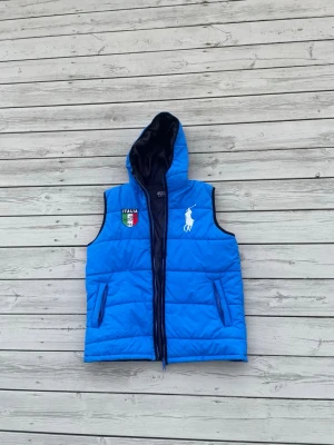 Blå dunväst från Polo Ralph Lauren - Snygg blå dunväst med huva från Polo Ralph Lauren. Västen har broderad logga på bröstet och ett Italia-märke med grönt, vitt och rött. Två dragkedjefickor och svart innerfoder. Perfekt för lager-på-lager och streetstyle.