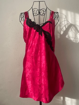 Rosa nattlinne från Vintage Victoria’s Secret - Snyggt nattlinne i starkt rosa satin med blommönster och bred svart spetsdetalj över bysten. Vintage Gold Label från VS. Storlek: S, passar 36-38 då VS Gold Label ofta är stor i storleken. Dockans storlek: 34.