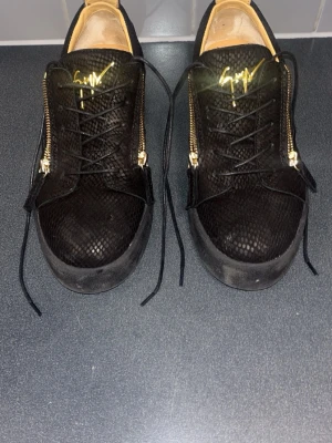 Svarta sneakers från Giuseppe Zanotti - Säljer ett par svarta sneakers från Giuseppe Zanotti med guldfärgade dragkedjor och logga. Skorna har en cool ormmönstrad textur i mocka och svarta snören. Insidan är beige och sulan är svart. Perfekta för dig som gillar exklusiv och unik stil. Det är Storlek 43 fast det passar 42