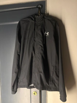 Svart vindjacka från Under Armour - Snygg svart vindjacka från Under Armour med huva och dragkedja framtill. Jackan har två sidofickor och ett vitt logotryck på bröstet. Tillverkad i lätt och vindtätt material, perfekt för blåsiga dagar. Enkel och stilren design som passar till det mesta.