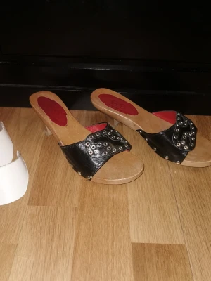 Svarta träklackade sandaler med nitar - Snygga sandaler med träklack och svart läderrem dekorerad med silvriga nitar. Röd logga på innersulan och öppen tå. Perfekta för dig som gillar en edgy och trendig stil. Är i fint använt skick, har några mindre defekter (se bild). Storlek 37 